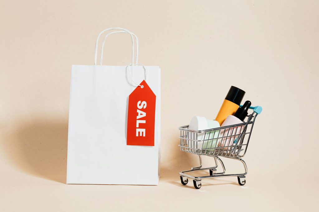 White shopping bag with red sale tag and mini shopping cart on beige background.